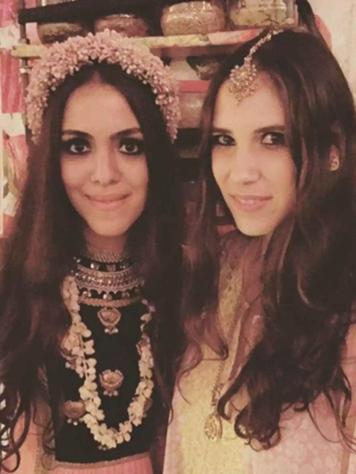 Dos mujeres jóvenes con cabello largo y suelto, vestidas con ropa tradicional y accesorios llamativos, posan juntas y sonríen frente a una estantería decorada. Dos mujeres jóvenes con cabello largo y suelto, vestidas con ropa tradicional y accesorios llamativos, posan juntas y sonríen frente a una estantería decorada.