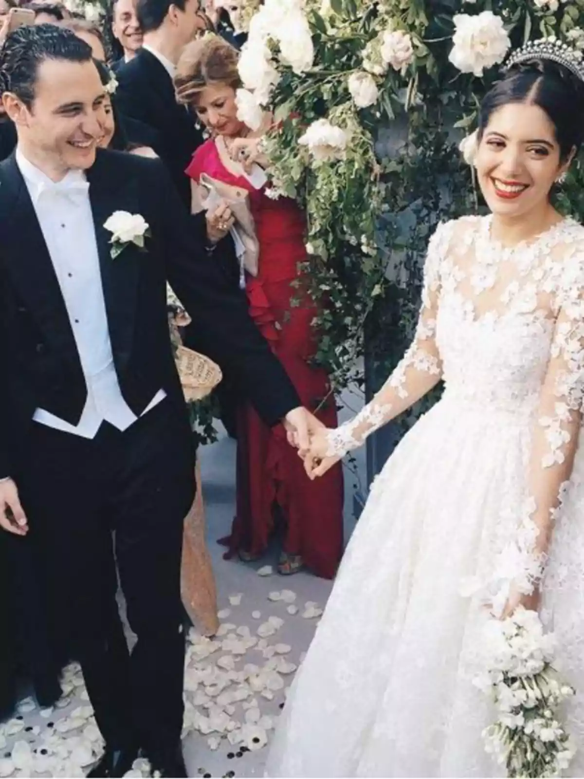 Pareja de recién casados sonríe mientras camina tomada de la mano entre pétalos de flores y decoración floral blanca Pareja de recién casados sonríe mientras camina tomada de la mano entre pétalos de flores y decoración floral blanca