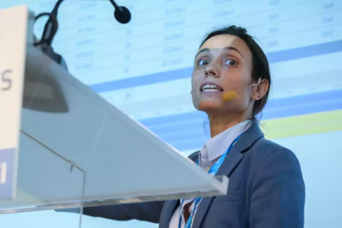 Mujer con micrófono de diadema dando una presentación frente a una pantalla con gráficos y datos. Mujer con micrófono de diadema dando una presentación frente a una pantalla con gráficos y datos.