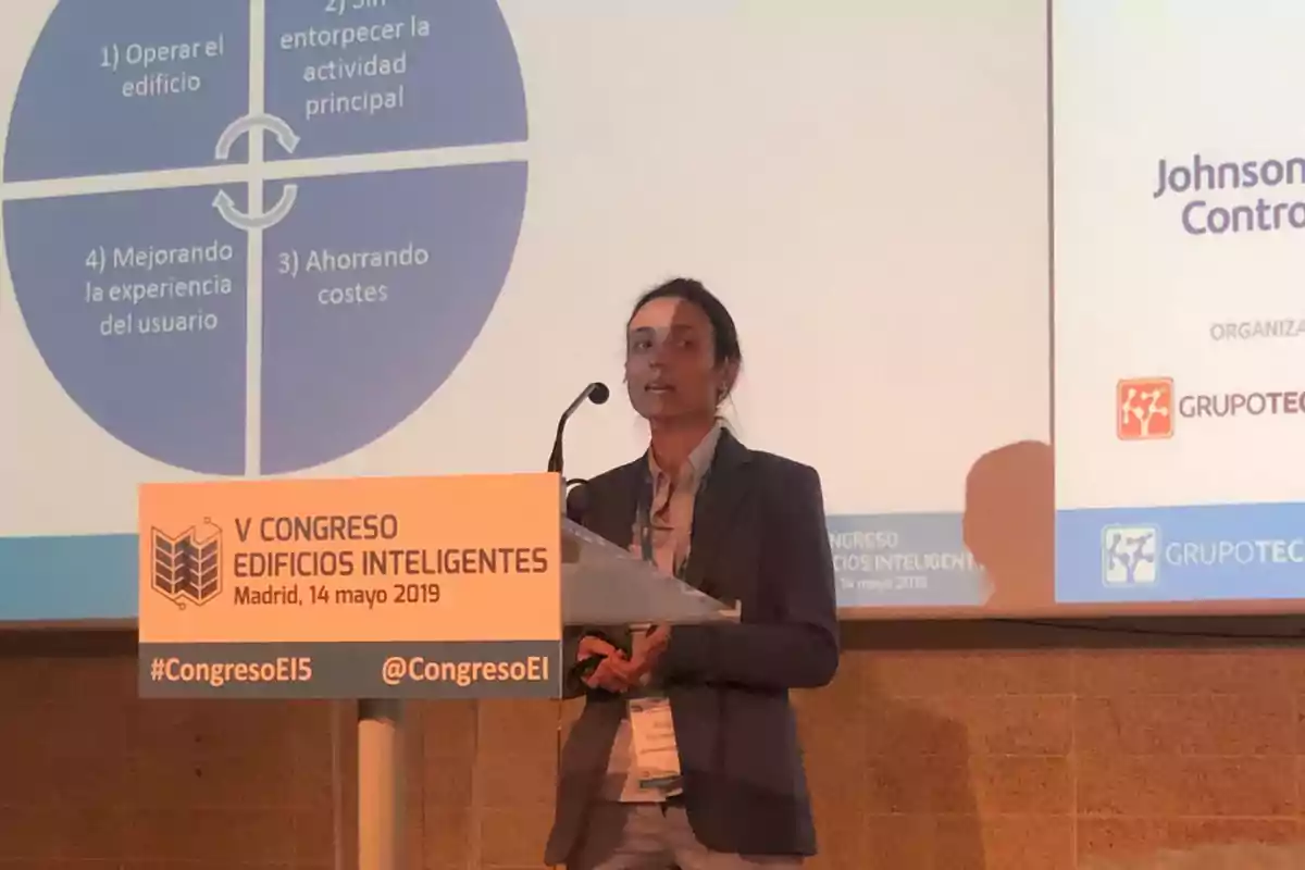 Mujer dando una presentación en el V Congreso Edificios Inteligentes en Madrid el 14 de mayo de 2019, con un gráfico proyectado detrás y un atril con el nombre del evento. Mujer dando una presentación en el V Congreso Edificios Inteligentes en Madrid el 14 de mayo de 2019, con un gráfico proyectado detrás y un atril con el nombre del evento.