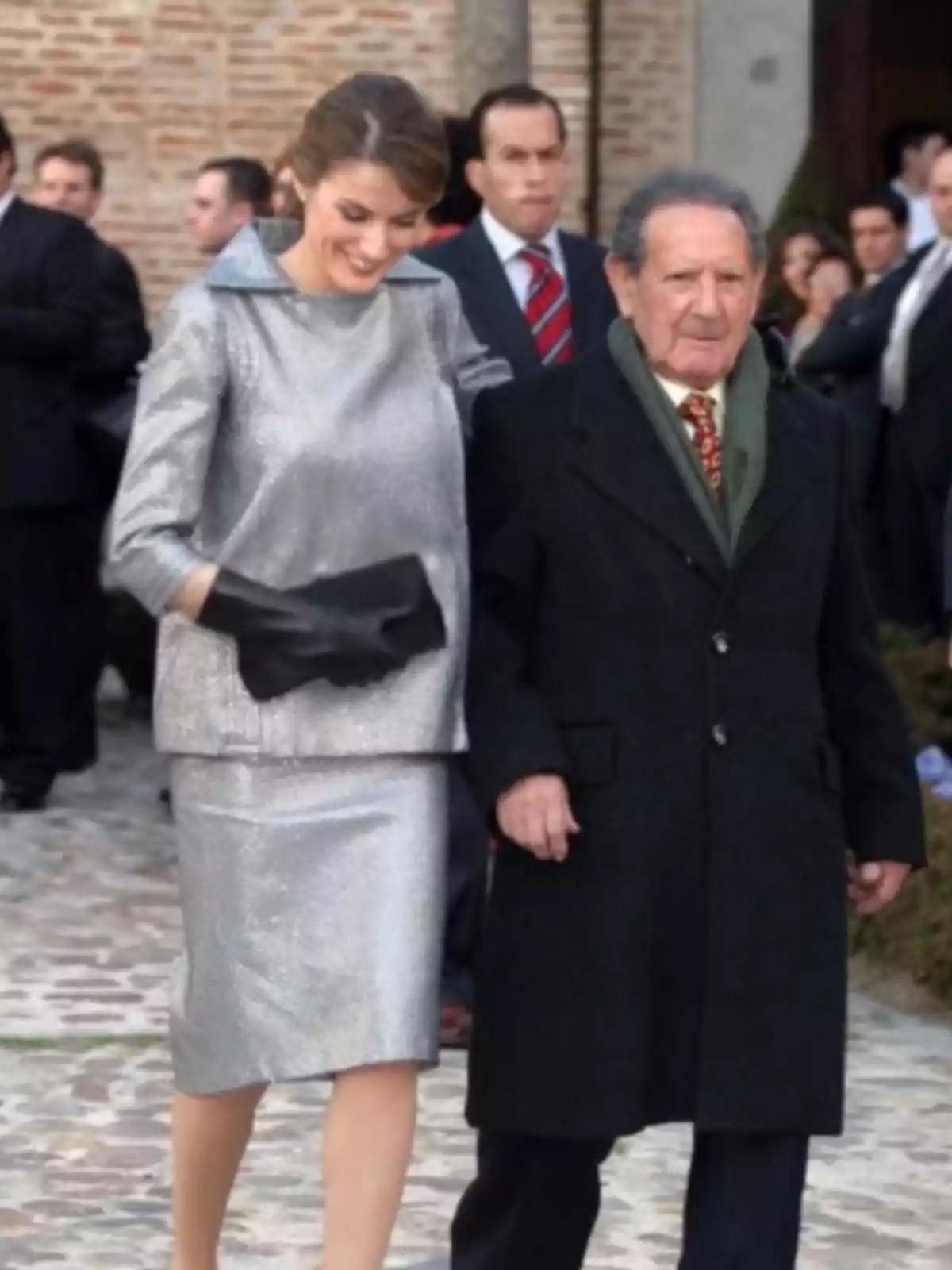 Una mujer vestida con un conjunto plateado camina del brazo de un hombre mayor con abrigo oscuro en un evento formal al aire libre rodeados de otras personas Una mujer vestida con un conjunto plateado camina del brazo de un hombre mayor con abrigo oscuro en un evento formal al aire libre rodeados de otras personas