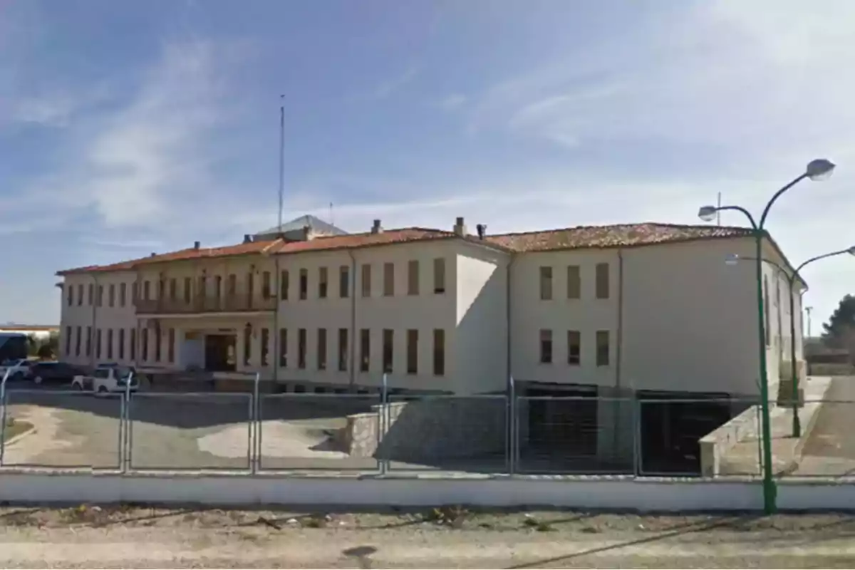 Edificio grande de dos pisos con techo de tejas rojas y muchas ventanas rodeado por una cerca metálica y con algunos coches estacionados al frente bajo un cielo despejado Edificio grande de dos pisos con techo de tejas rojas y muchas ventanas rodeado por una cerca metálica y con algunos coches estacionados al frente bajo un cielo despejado