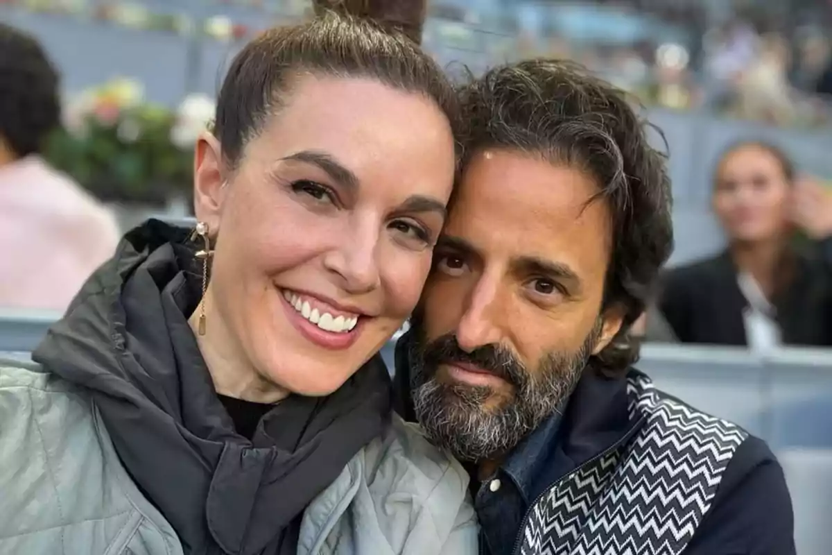 Una pareja sonríe y posa para una selfie en un lugar público con personas al fondo. Una pareja sonríe y posa para una selfie en un lugar público con personas al fondo.