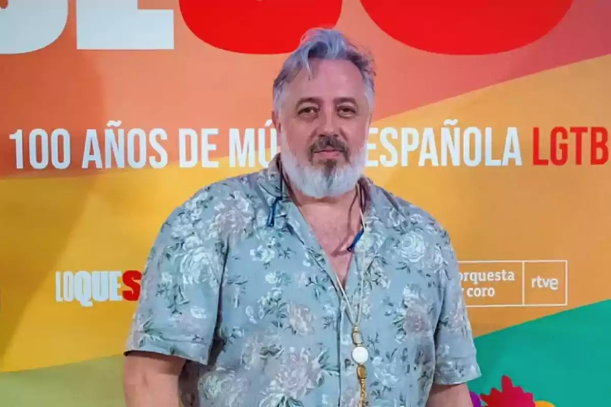 Hombre de cabello y barba canosos con camisa de flores posando frente a un fondo colorido con texto sobre música española y LGTB Hombre de cabello y barba canosos con camisa de flores posando frente a un fondo colorido con texto sobre música española y LGTB