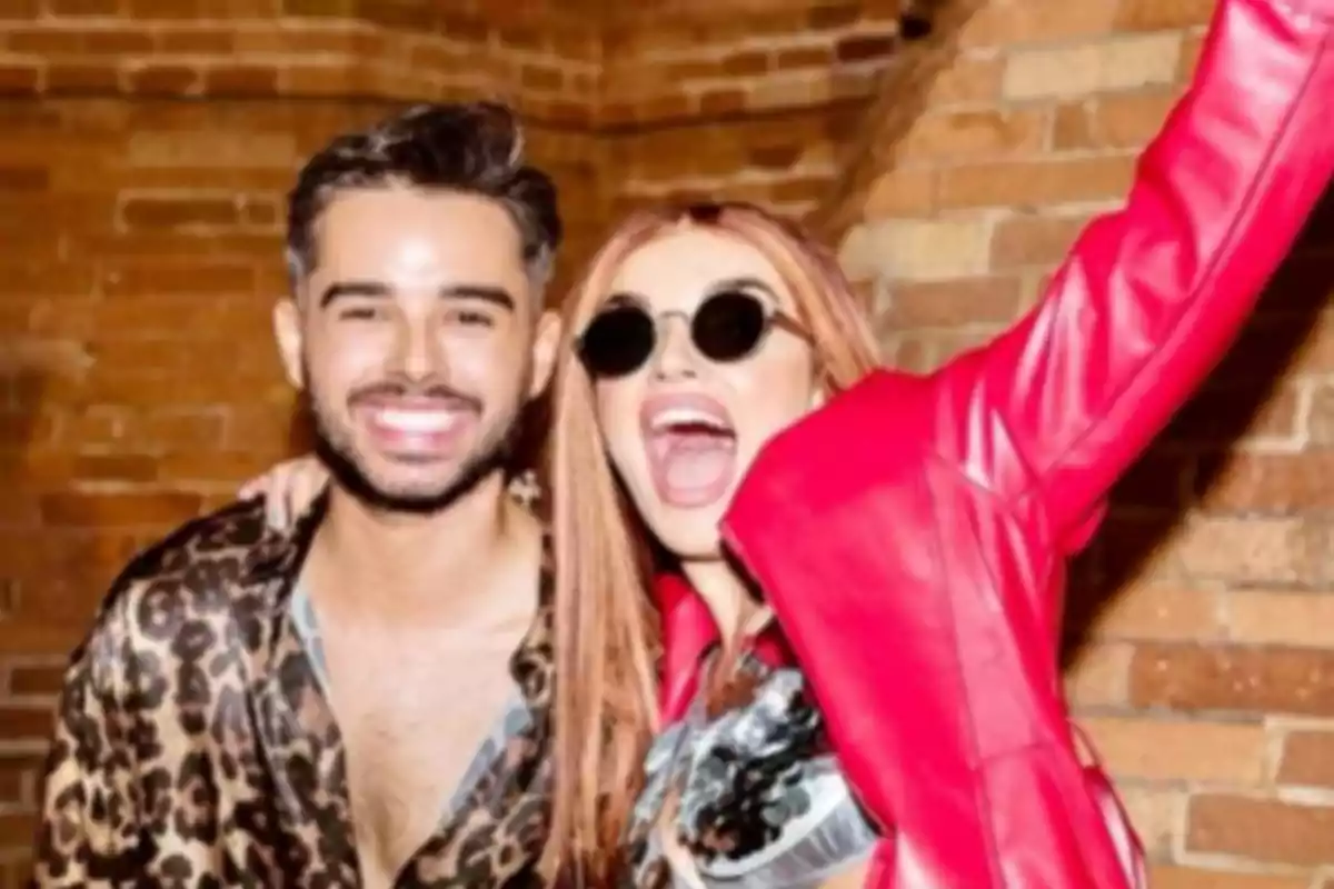 Dos personas sonríen y posan felices frente a una pared de ladrillos, una de ellas lleva gafas oscuras y chaqueta roja Dos personas sonríen y posan felices frente a una pared de ladrillos, una de ellas lleva gafas oscuras y chaqueta roja