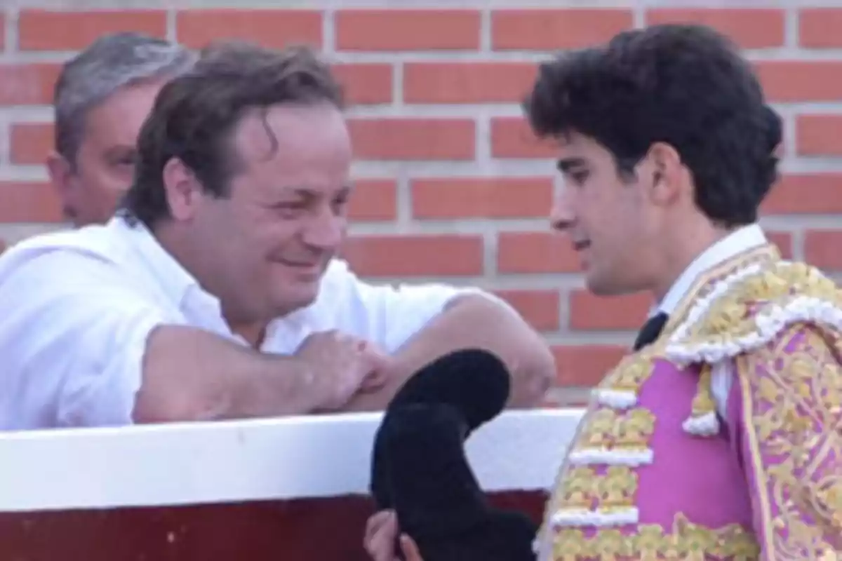 Un hombre vestido de blanco sonríe y conversa con un torero que lleva un traje de luces color rosa y dorado frente a una pared de ladrillos Un hombre vestido de blanco sonríe y conversa con un torero que lleva un traje de luces color rosa y dorado frente a una pared de ladrillos