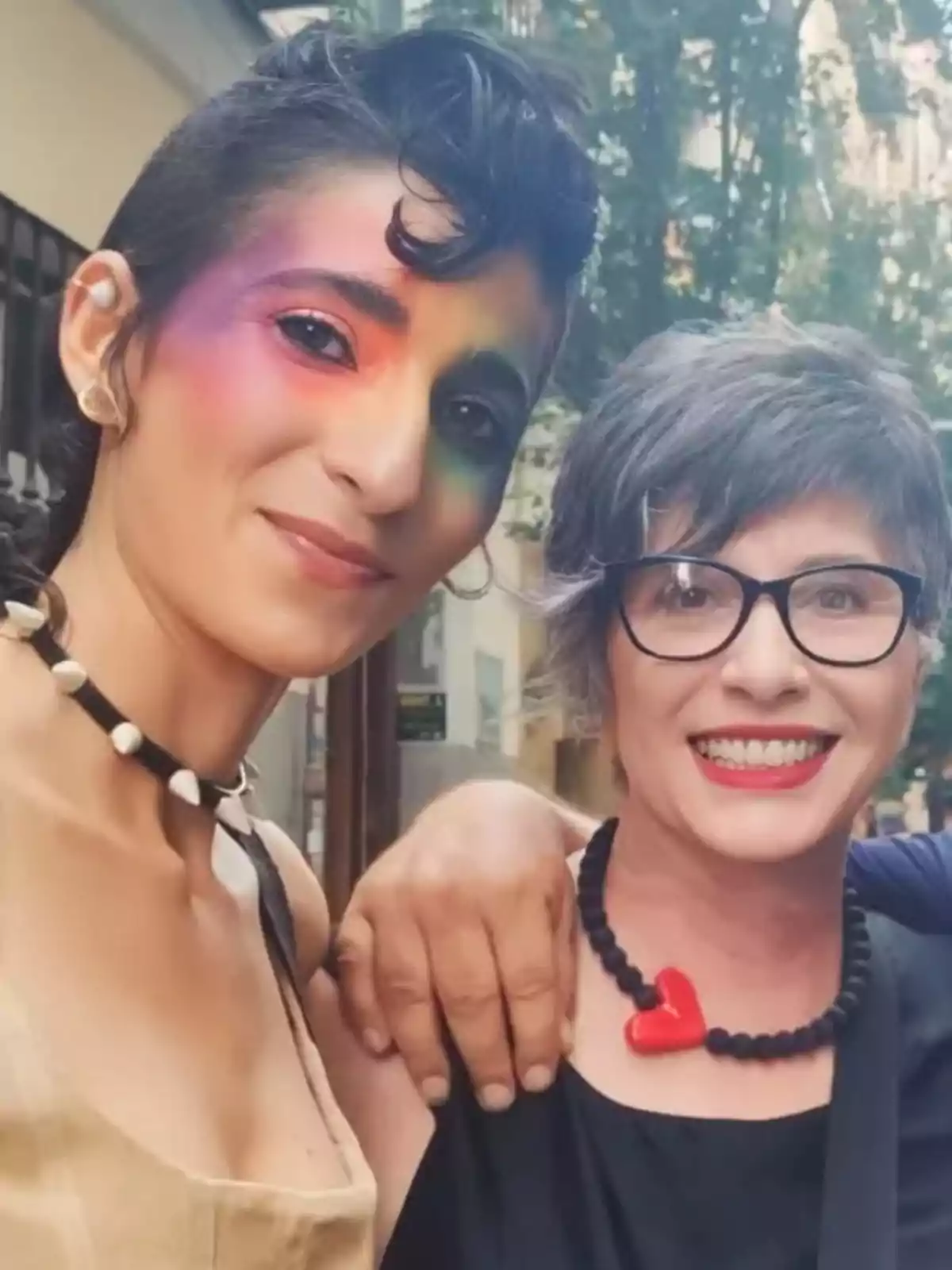 Dos personas sonríen a la cámara, una lleva maquillaje colorido en los ojos y la otra usa gafas y un collar con un corazón rojo, ambas están al aire libre. Dos personas sonríen a la cámara, una lleva maquillaje colorido en los ojos y la otra usa gafas y un collar con un corazón rojo, ambas están al aire libre.