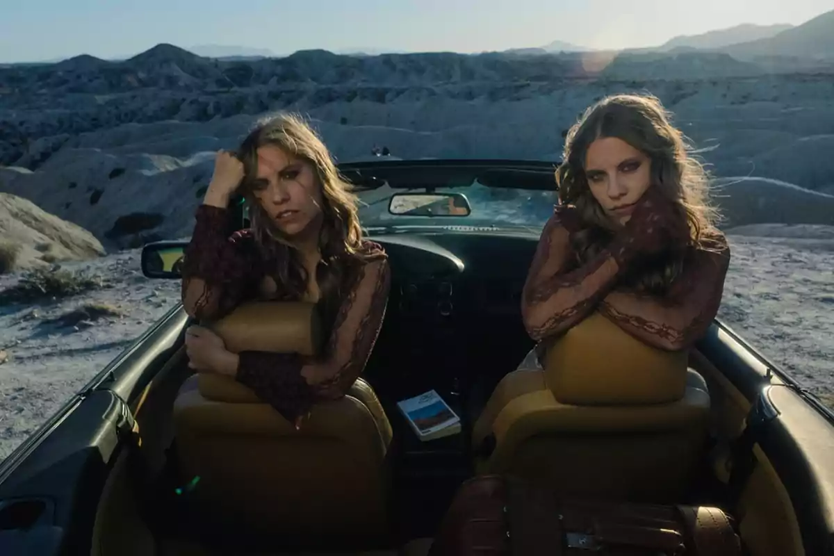 Dos mujeres con expresión seria sentadas en un auto descapotable en medio de un paisaje árido y montañoso