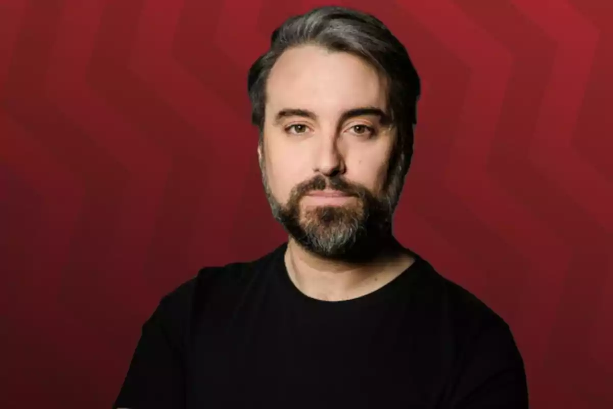 Hombre de cabello y barba oscura con algunas canas, vistiendo camiseta negra y posando frente a un fondo rojo con patrón geométrico