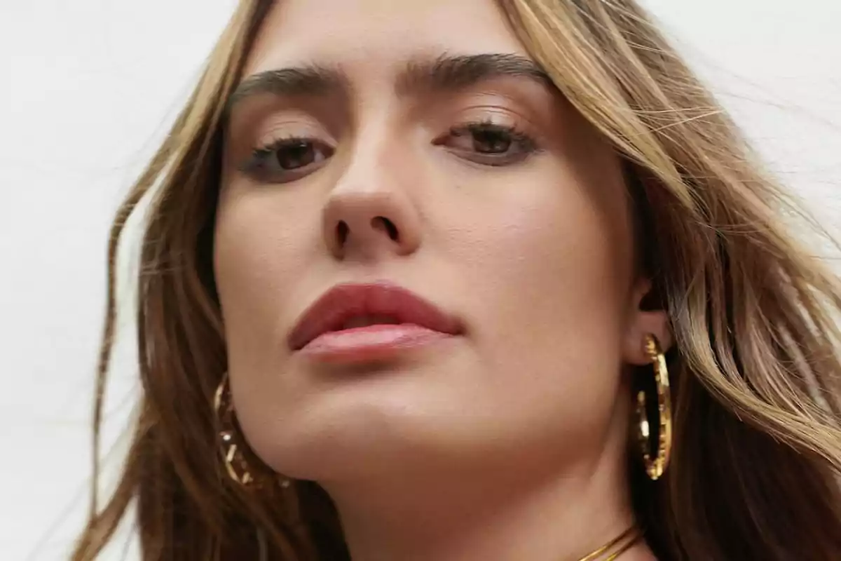 Mujer de cabello castaño claro con ondas, usando aretes grandes dorados y un collar dorado, mirando hacia la cámara con expresión seria