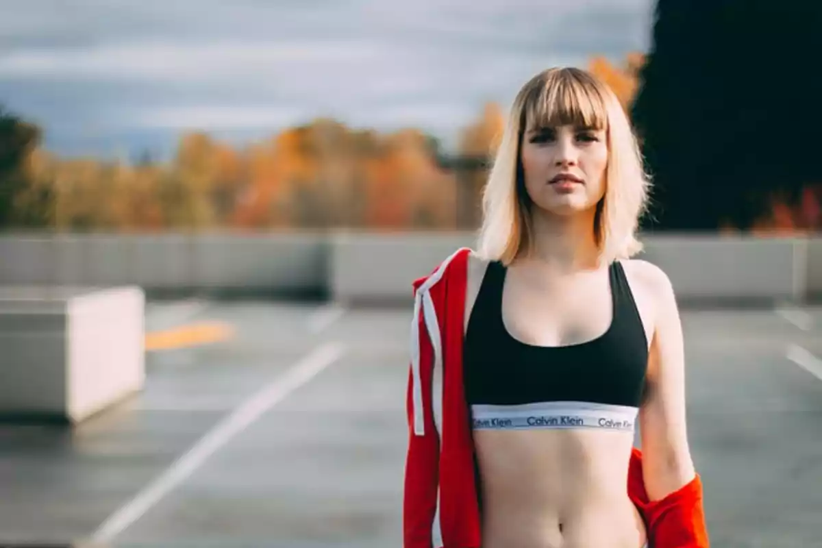 Mujer joven con cabello rubio y flequillo usando top deportivo negro y chaqueta roja en un estacionamiento al aire libre con árboles de fondo