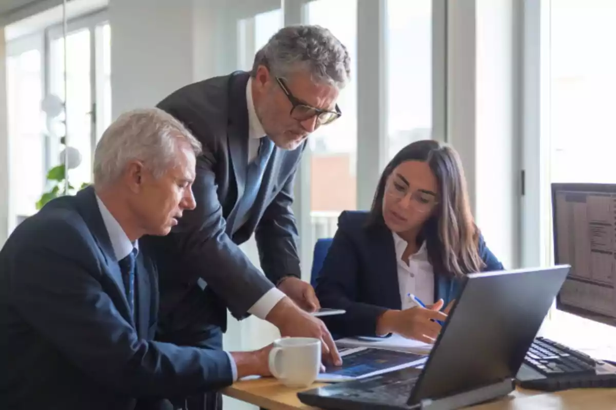 Tres personas vestidas de traje trabajando juntas en una oficina, revisando documentos y usando una computadora portátil