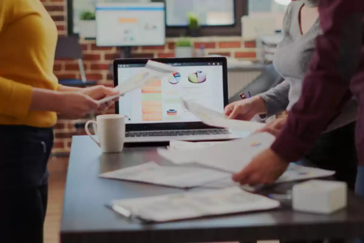 Personas revisando documentos y gráficos en una mesa de oficina con una laptop encendida al fondo