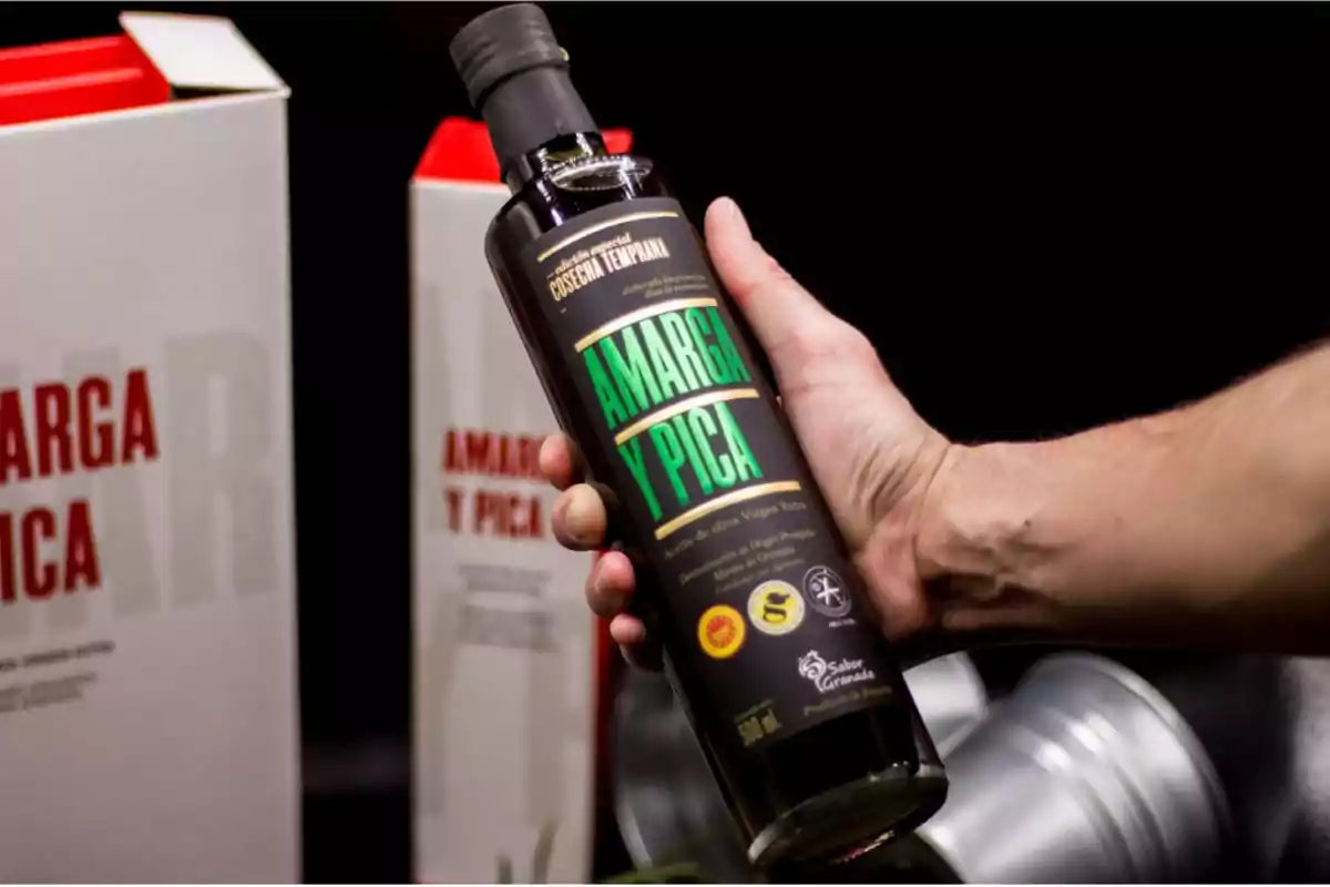 Persona sosteniendo una botella de aceite de oliva virgen extra llamada Amarga y Pica con cajas del mismo producto al fondo sobre un fondo negro
