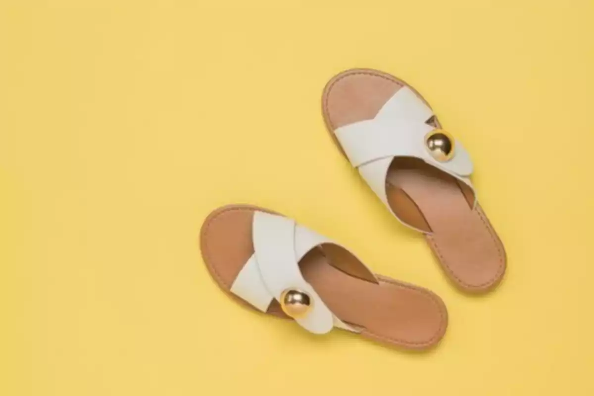 Sandalias planas blancas con detalle dorado sobre fondo amarillo