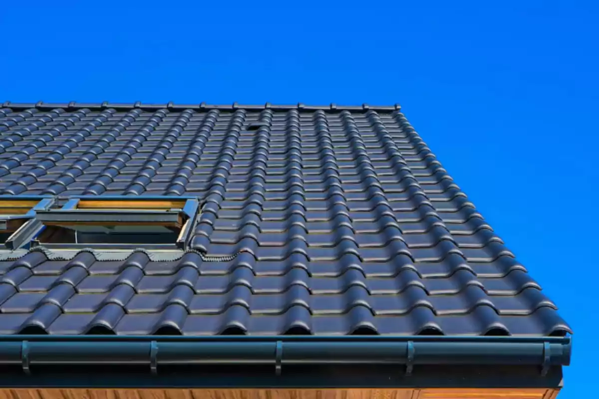 Tejado de tejas cerámicas negras con una ventana abierta bajo un cielo azul despejado