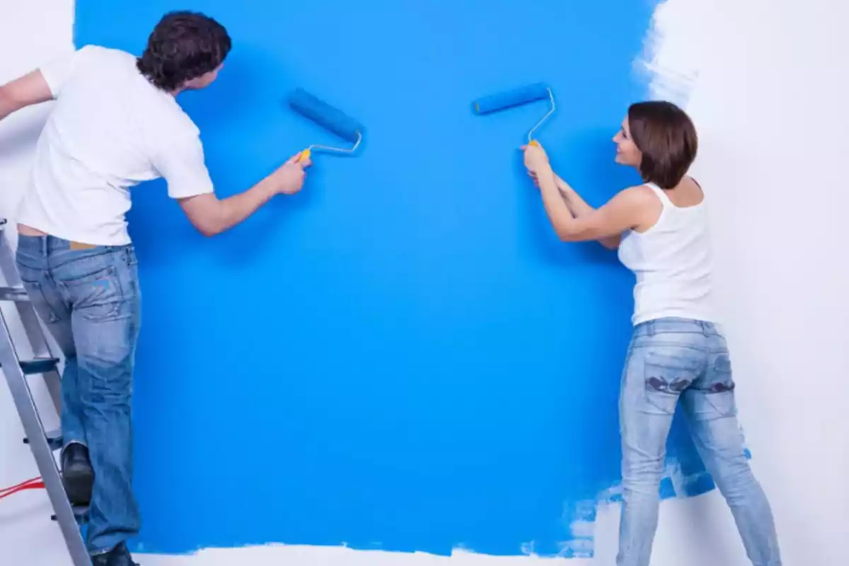 Dos personas pintan una pared de azul usando rodillos mientras sonríen y trabajan juntas