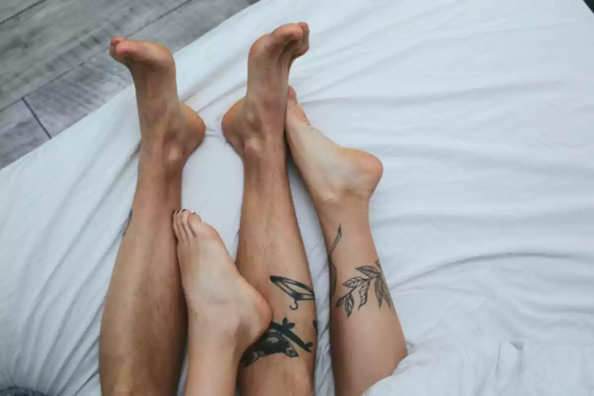 Dos personas con tatuajes en las piernas están recostadas juntas sobre una cama con sábanas blancas