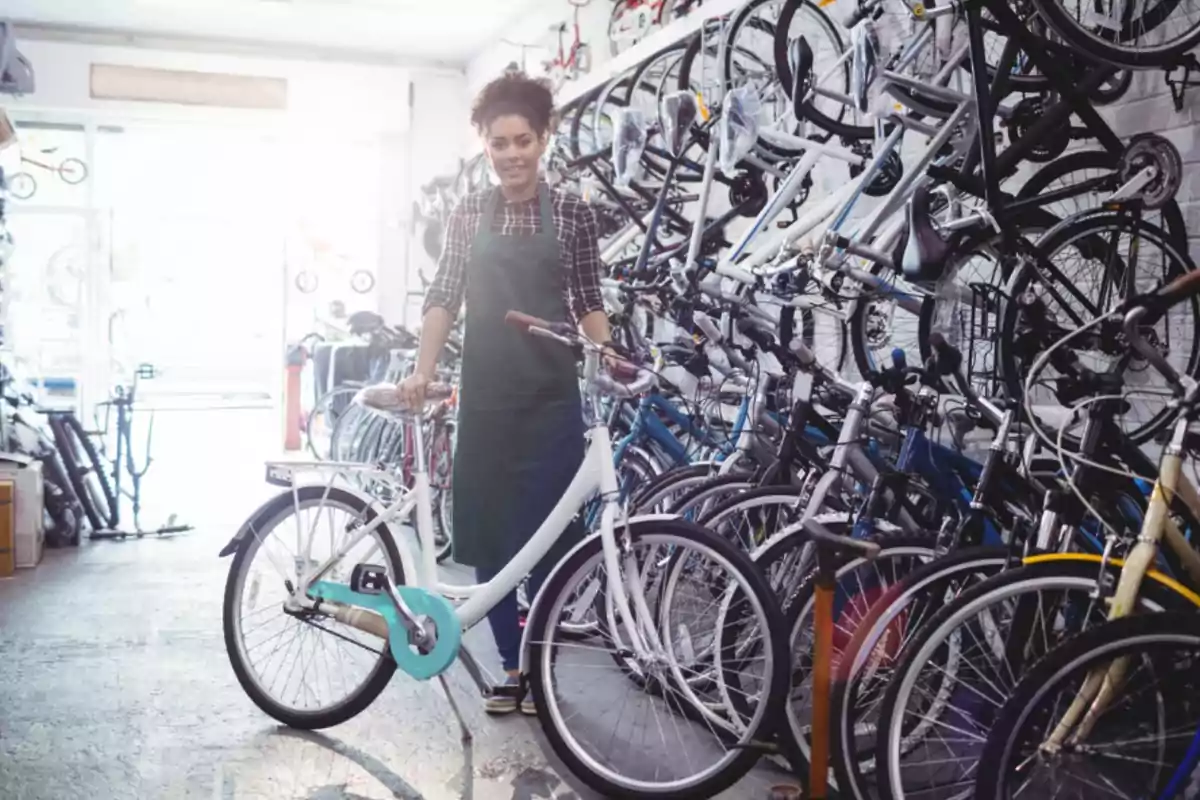 Mujer joven con delantal sonríe mientras sostiene una bicicleta blanca en el interior de una tienda llena de bicicletas exhibidas en las paredes y el suelo