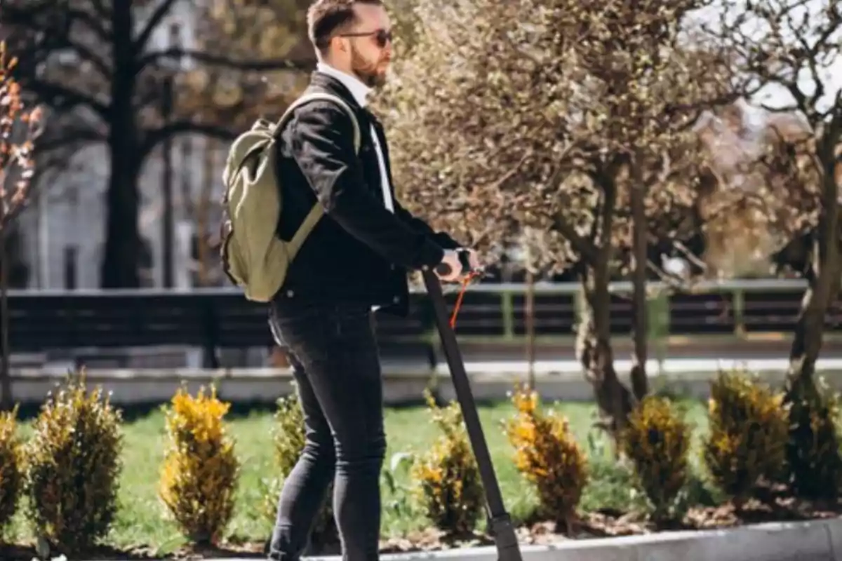 Hombre joven con barba y gafas de sol montando un patinete eléctrico en un parque mientras lleva una mochila verde