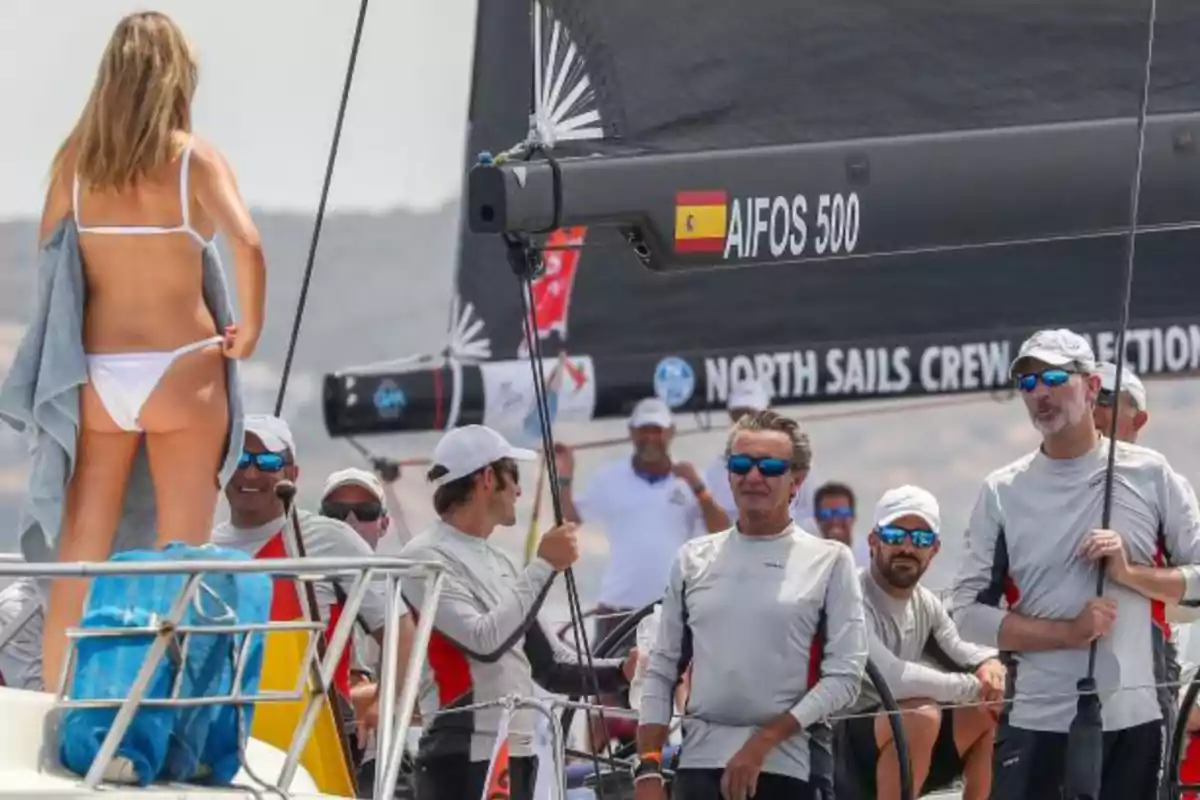 A woman in a white bikini seen from behind and several men in sportswear and sunglasses on a sailboat with a sail bearing the inscription AIFOS 500 and a Spanish flag. A woman in a white bikini seen from behind and several men in sportswear and sunglasses on a sailboat with a sail bearing the inscription AIFOS 500 and a Spanish flag.