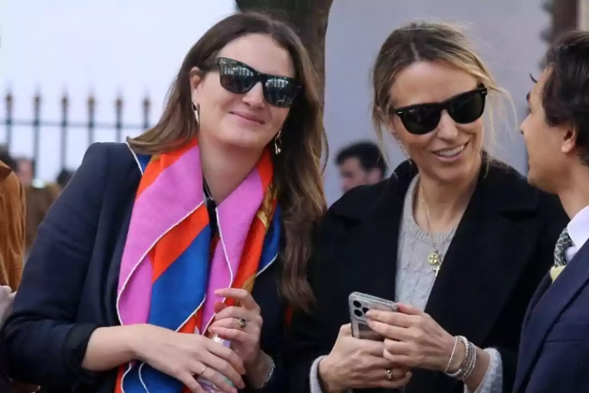 Dos mujeres con gafas de sol sonríen mientras conversan con un hombre, una de ellas lleva un pañuelo colorido y la otra sostiene un teléfono móvil Dos mujeres con gafas de sol sonríen mientras conversan con un hombre, una de ellas lleva un pañuelo colorido y la otra sostiene un teléfono móvil