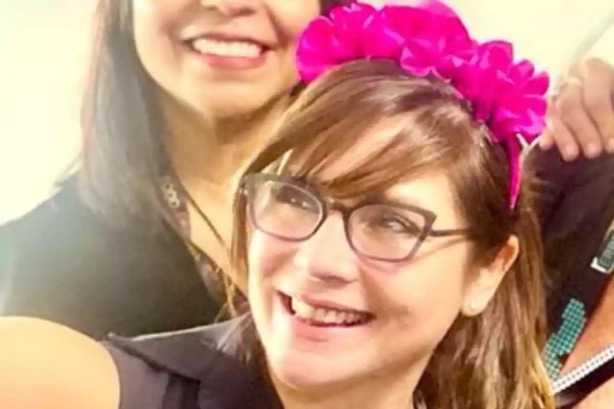 Dos mujeres sonríen mientras una de ellas lleva una diadema de flores rosas y gafas Dos mujeres sonríen mientras una de ellas lleva una diadema de flores rosas y gafas