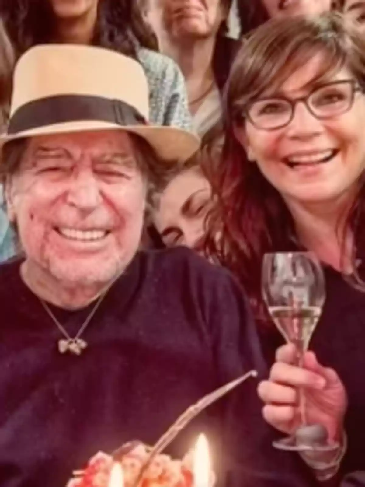 Un hombre mayor con sombrero sonríe junto a una mujer con gafas que sostiene una copa de vino, rodeados de más personas en un ambiente festivo Un hombre mayor con sombrero sonríe junto a una mujer con gafas que sostiene una copa de vino, rodeados de más personas en un ambiente festivo