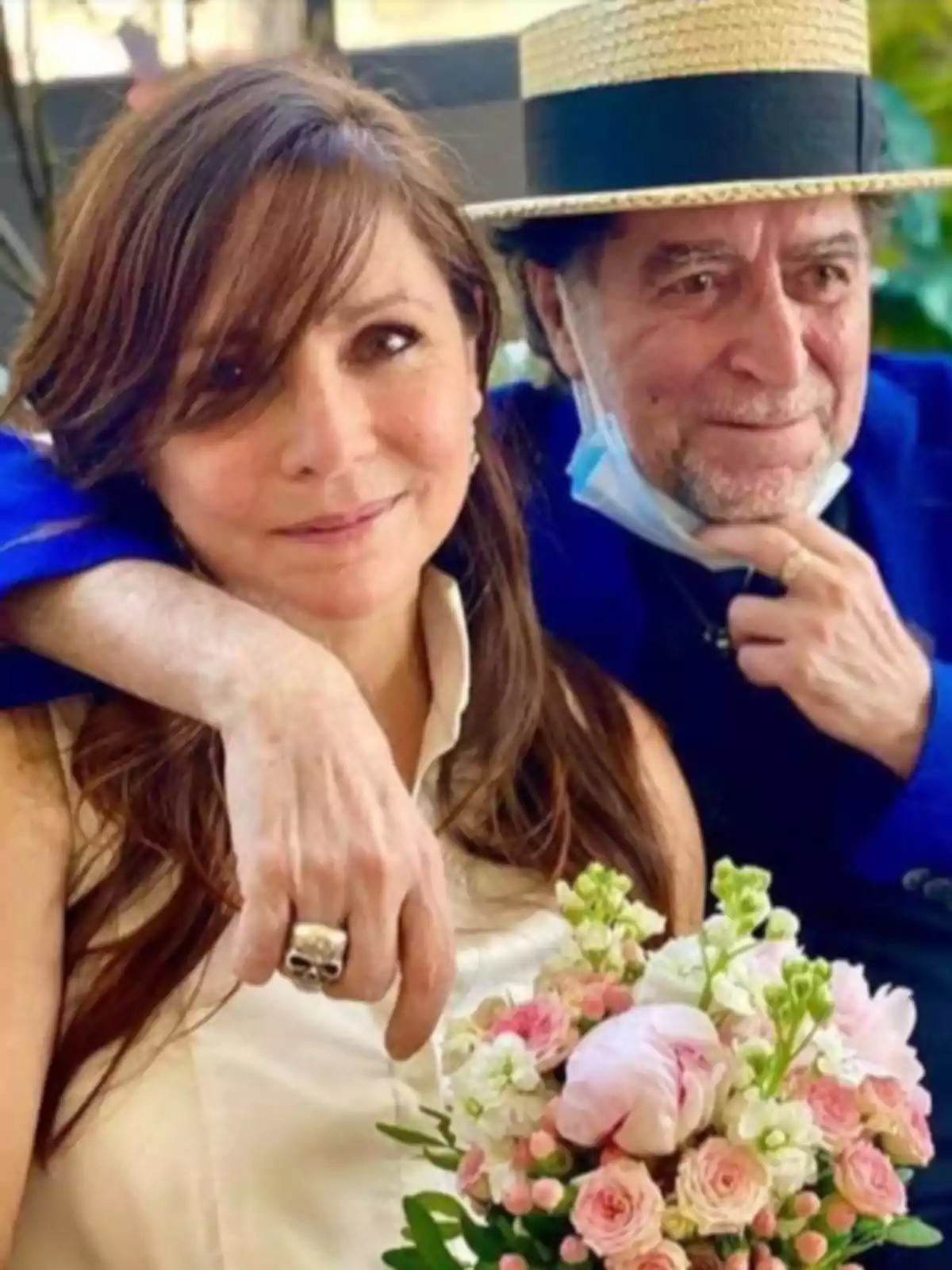 Una mujer sonriente sostiene un ramo de flores mientras un hombre con sombrero y mascarilla bajada la abraza por el hombro Una mujer sonriente sostiene un ramo de flores mientras un hombre con sombrero y mascarilla bajada la abraza por el hombro