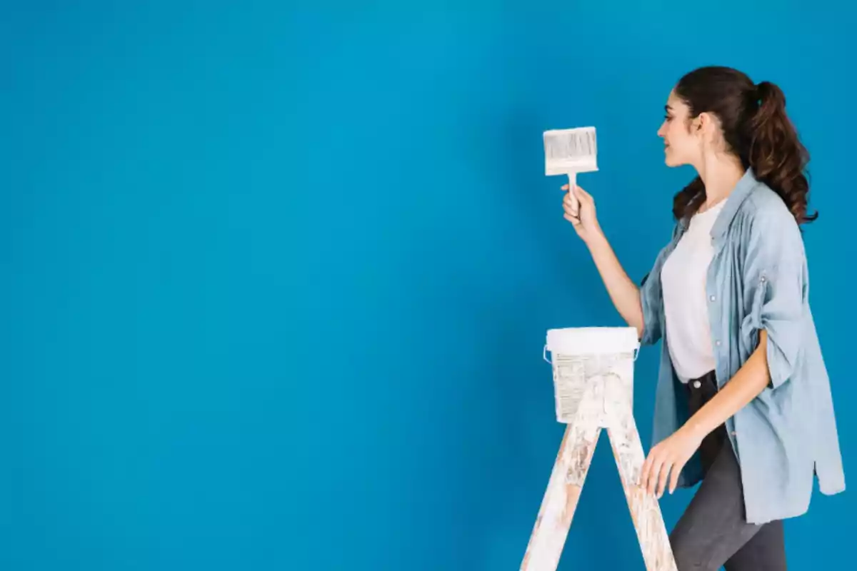 Mujer joven pintando una pared azul mientras sostiene una brocha y se apoya en una escalera