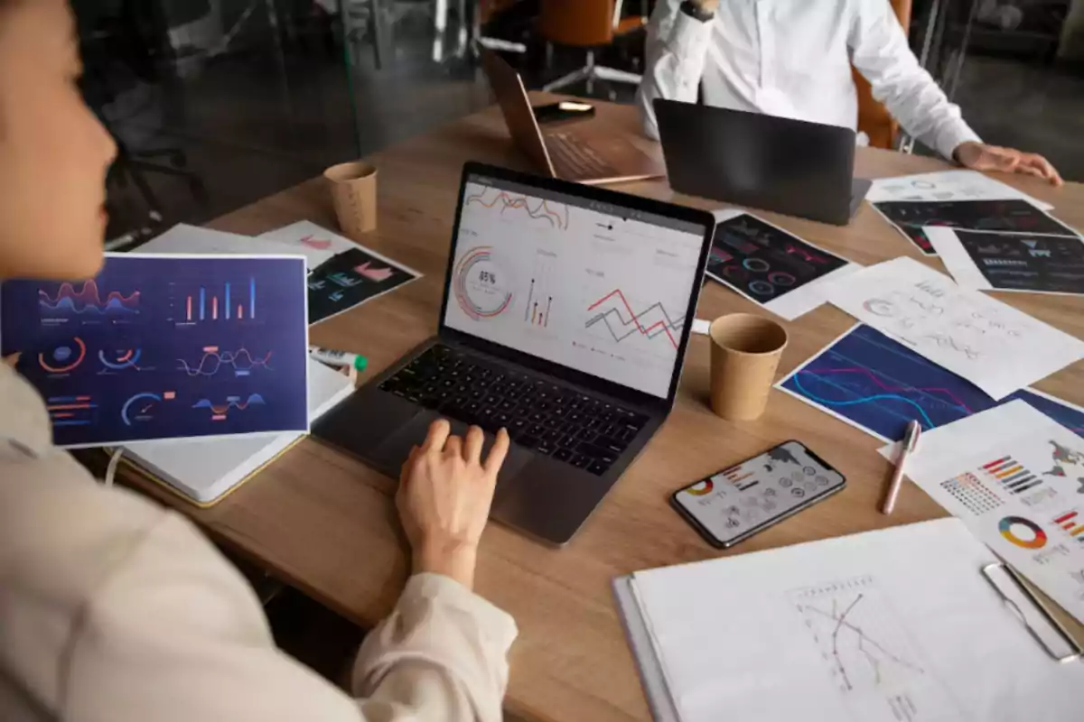 Personas trabajando en una mesa con computadoras portátiles y gráficos de datos impresos y digitales