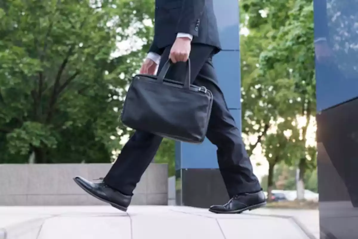 Hombre de negocios caminando con un maletín negro en la mano en un entorno urbano con árboles al fondo