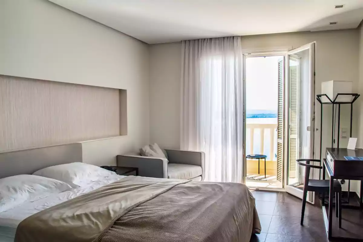 Habitación de hotel moderna y luminosa con cama deshecha, sillón, escritorio y balcón con vista al mar