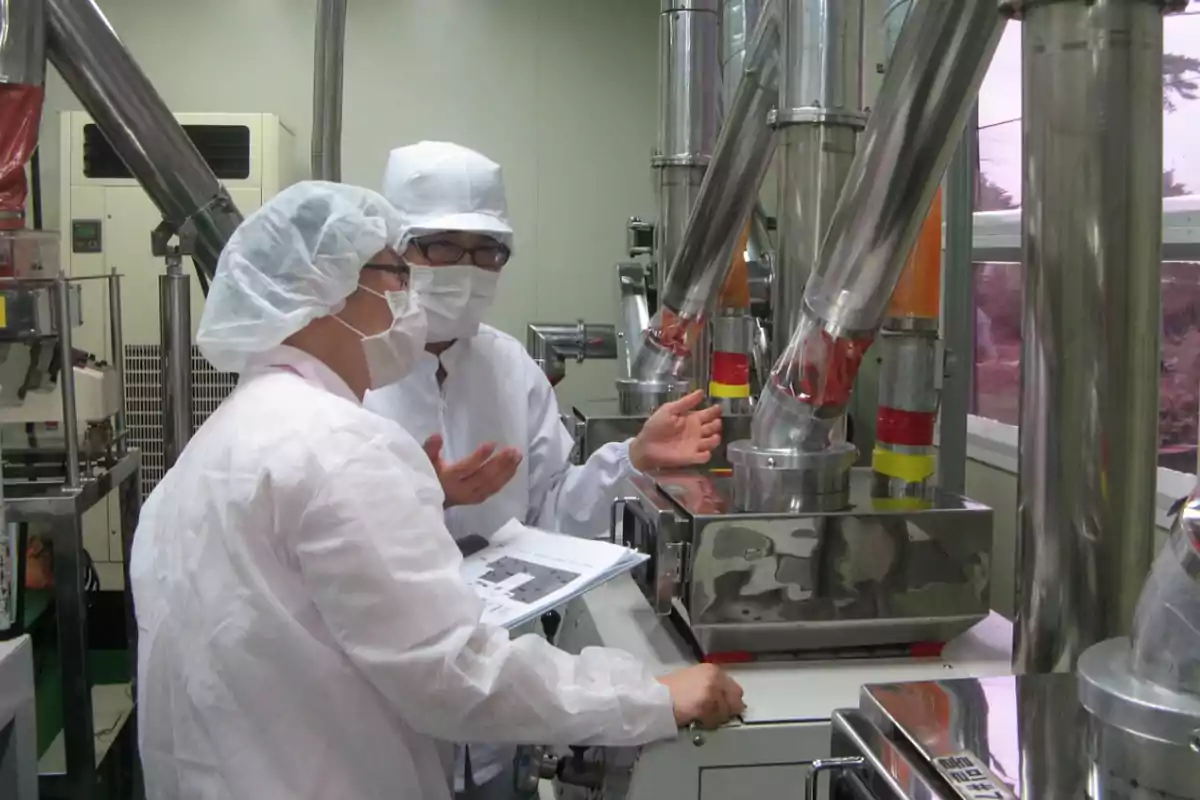 Dos personas con bata y mascarilla trabajan en un laboratorio industrial rodeados de maquinaria de acero inoxidable