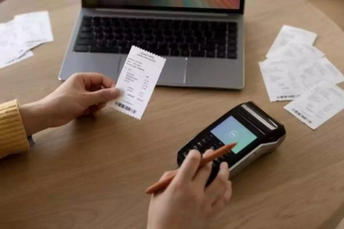 Persona revisando un recibo junto a una terminal de pago y una computadora portátil sobre una mesa