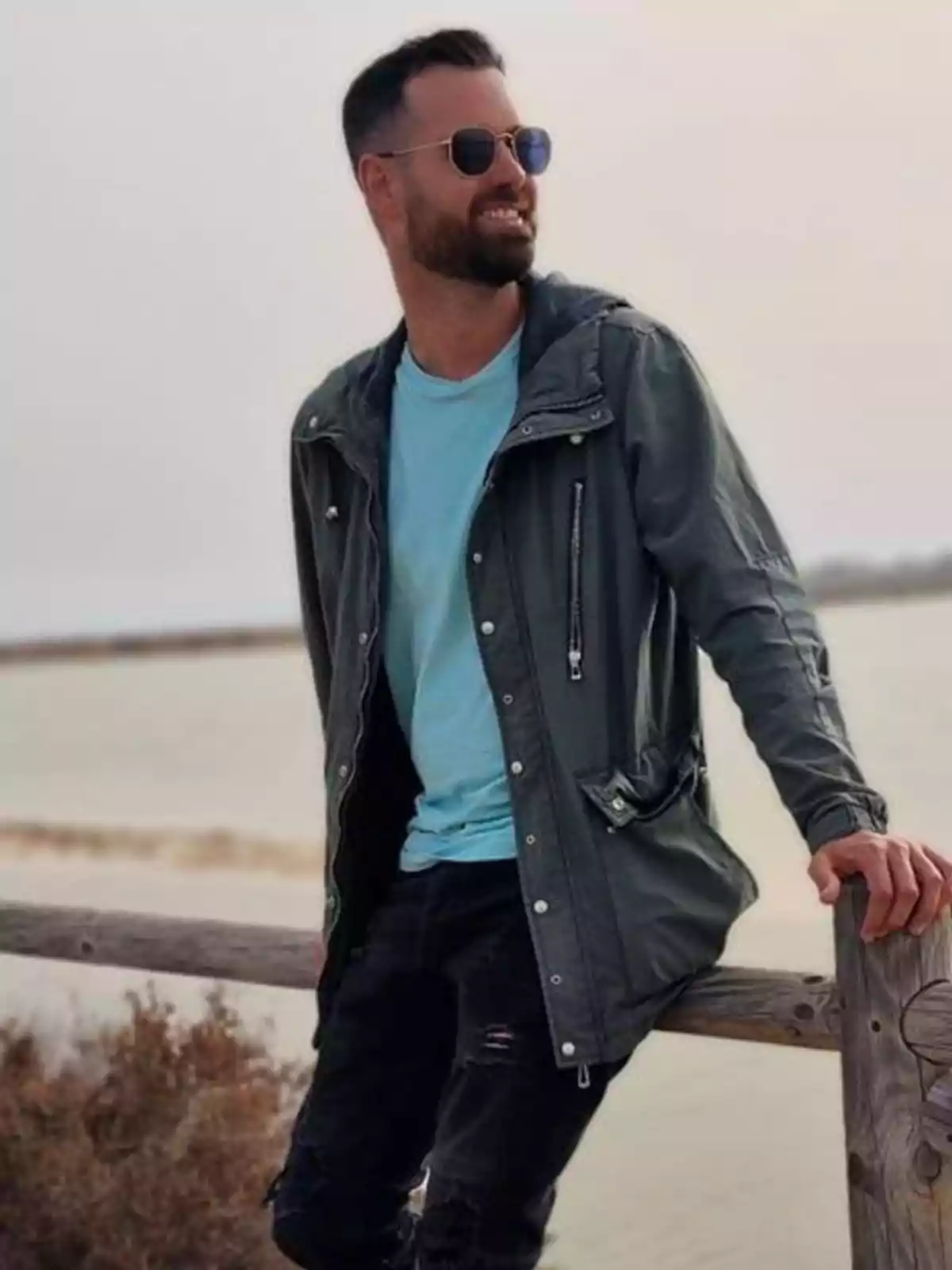 Hombre con barba y gafas de sol apoyado en una cerca de madera junto a un lago, vistiendo chaqueta verde y camiseta celeste, sonriendo y mirando hacia un lado. Hombre con barba y gafas de sol apoyado en una cerca de madera junto a un lago, vistiendo chaqueta verde y camiseta celeste, sonriendo y mirando hacia un lado.