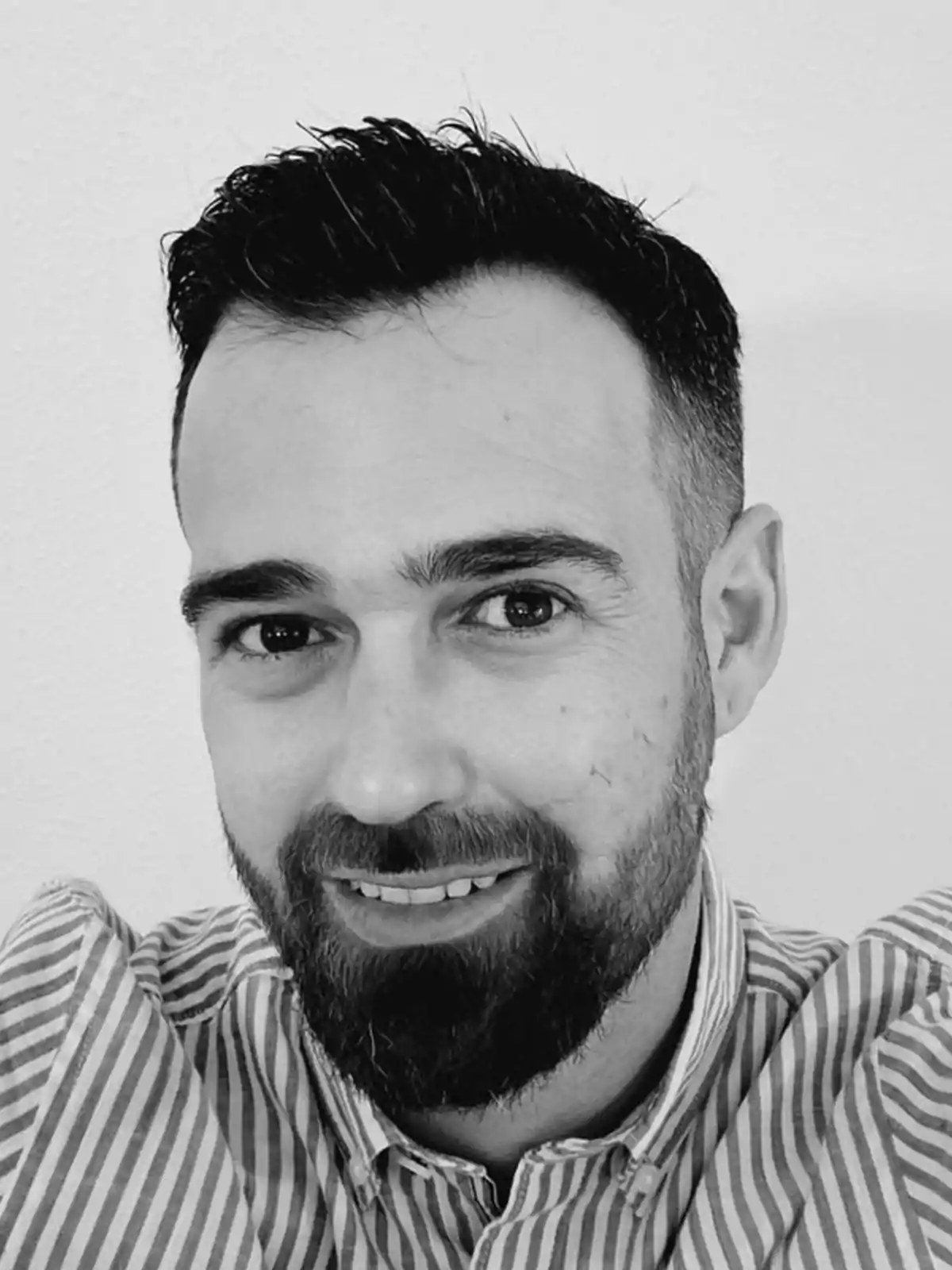 Hombre joven con barba y cabello corto, sonriendo y vistiendo una camisa de rayas, en una foto en blanco y negro Hombre joven con barba y cabello corto, sonriendo y vistiendo una camisa de rayas, en una foto en blanco y negro