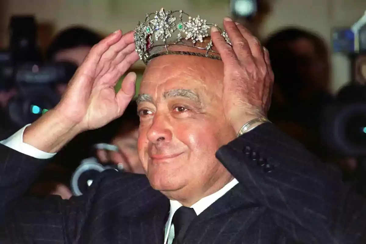 Hombre mayor con traje oscuro colocándose una corona de joyas en la cabeza mientras sonríe frente a cámaras Hombre mayor con traje oscuro colocándose una corona de joyas en la cabeza mientras sonríe frente a cámaras