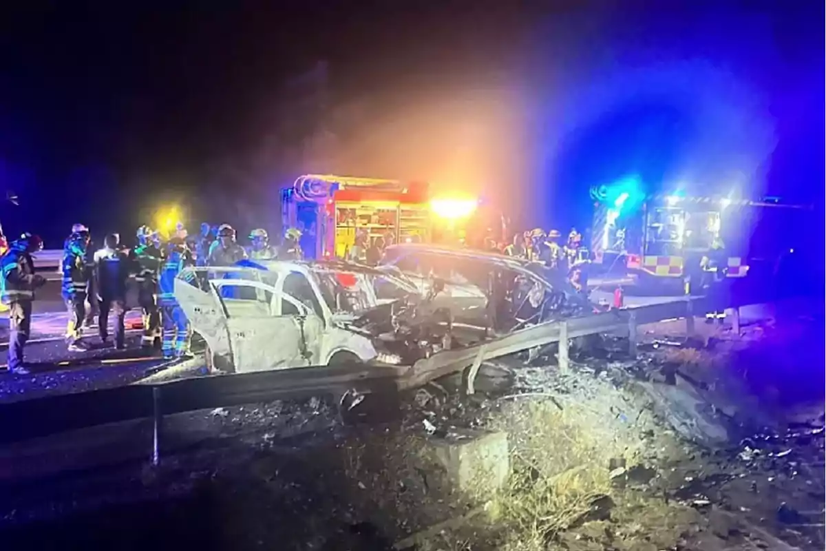 Accidente automovilístico nocturno con varios bomberos y vehículos de emergencia atendiendo la escena donde dos autos chocaron y resultaron dañados junto a una barrera metálica