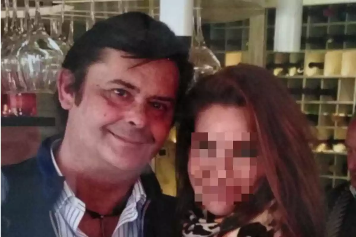 Un hombre sonriente posa junto a una mujer cuyo rostro está pixelado en un ambiente de restaurante elegante con copas colgadas y botellas al fondo Un hombre sonriente posa junto a una mujer cuyo rostro está pixelado en un ambiente de restaurante elegante con copas colgadas y botellas al fondo