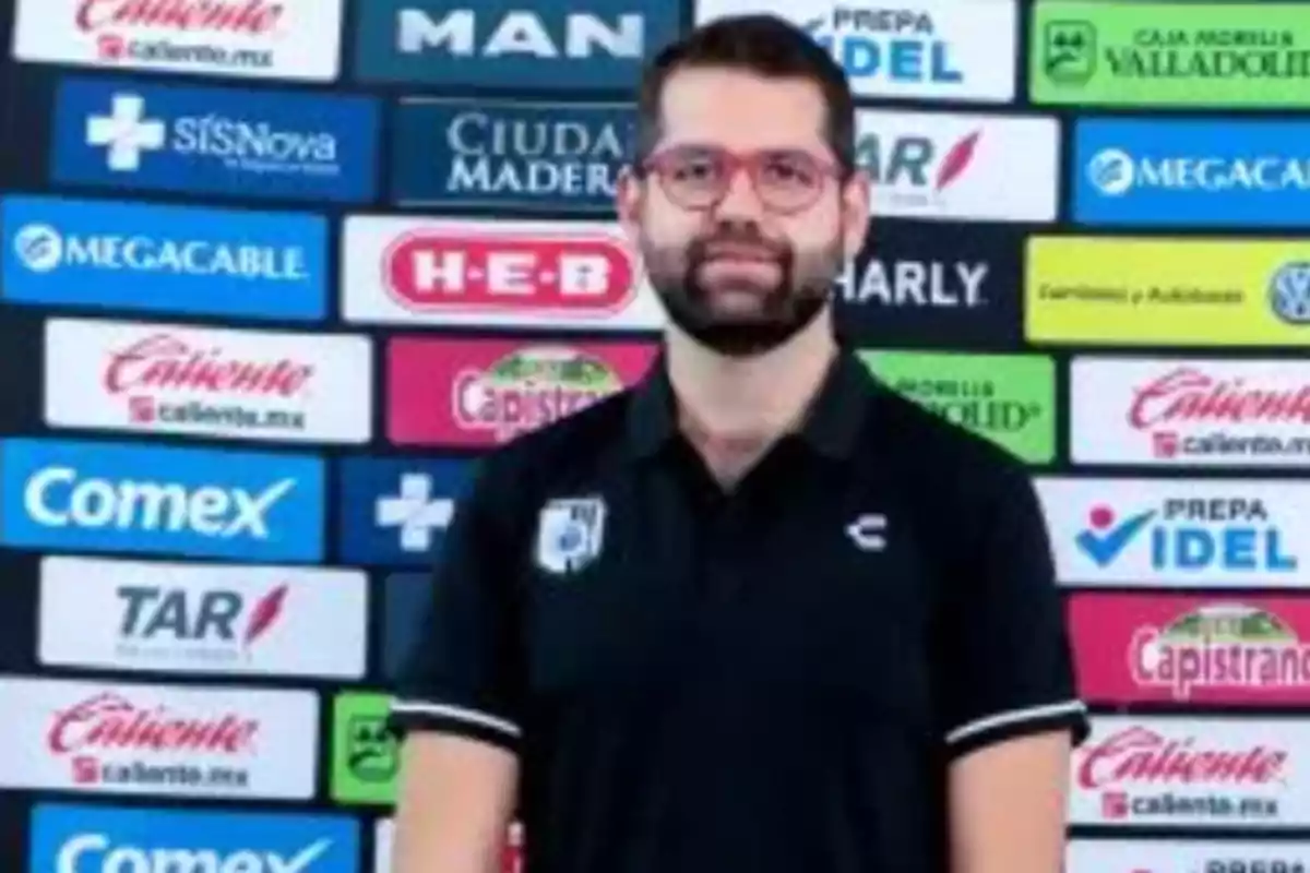 Hombre con barba y gafas de pasta posando frente a un fondo de logotipos de patrocinadores deportivos Hombre con barba y gafas de pasta posando frente a un fondo de logotipos de patrocinadores deportivos
