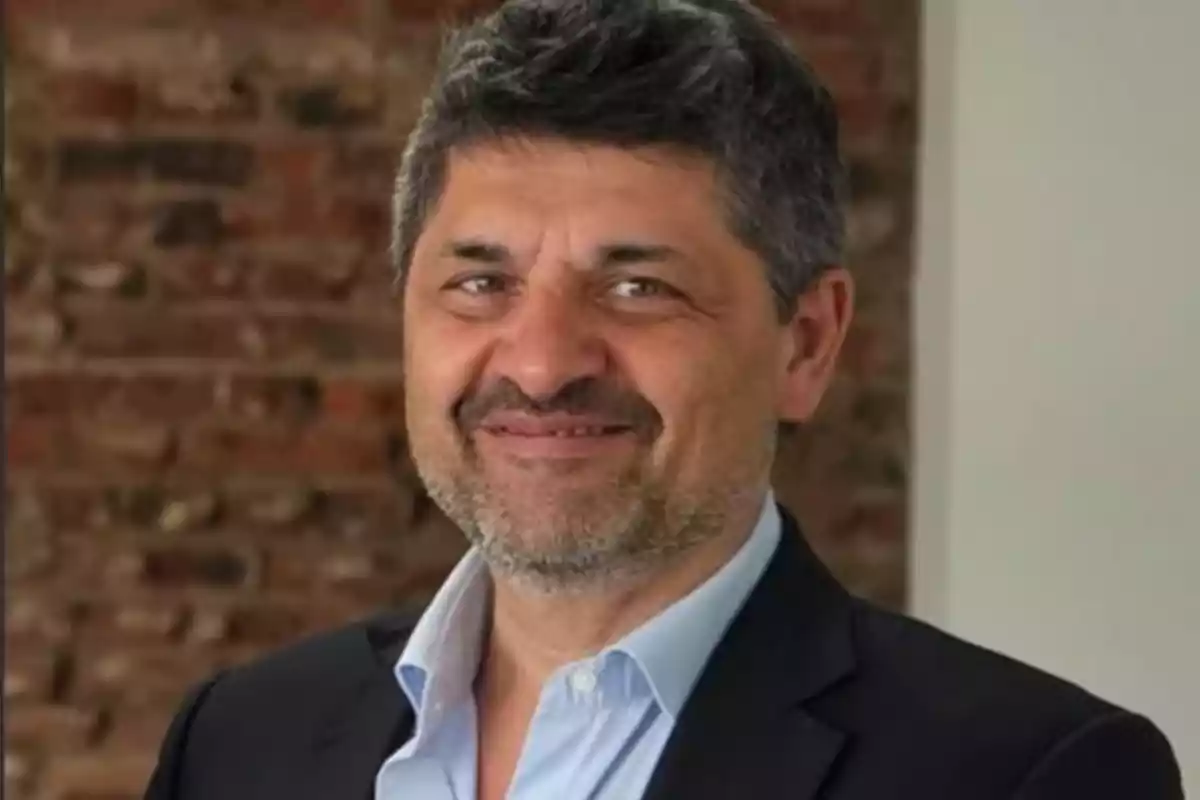 Hombre de cabello canoso y barba corta con saco oscuro y camisa celeste sonriendo frente a una pared de ladrillos Hombre de cabello canoso y barba corta con saco oscuro y camisa celeste sonriendo frente a una pared de ladrillos