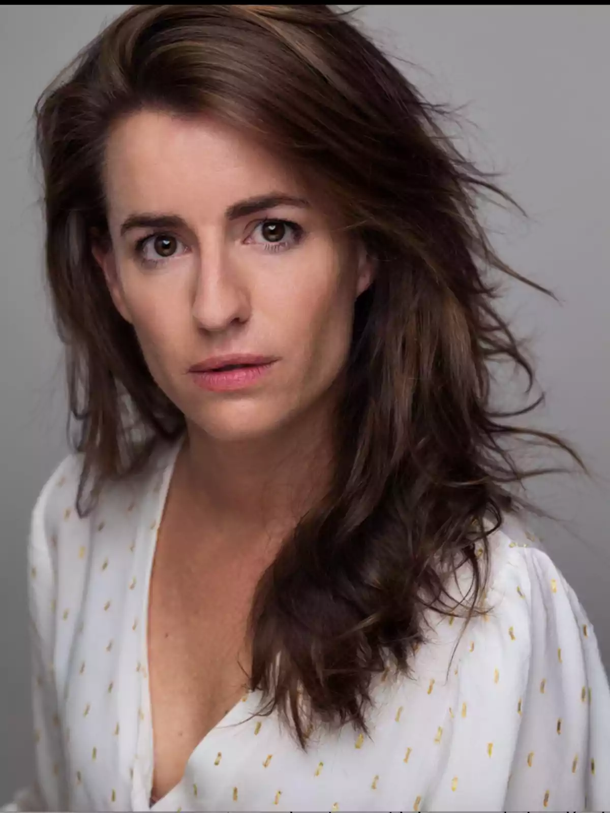 Mujer de cabello castaño largo y suelto con expresión seria, vestida con blusa blanca de detalles dorados, fondo gris neutro Mujer de cabello castaño largo y suelto con expresión seria, vestida con blusa blanca de detalles dorados, fondo gris neutro