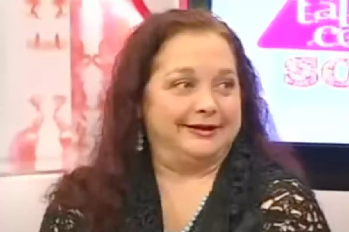 Mujer de cabello largo y oscuro con expresión sonriente sentada en un set de televisión con fondo colorido Mujer de cabello largo y oscuro con expresión sonriente sentada en un set de televisión con fondo colorido