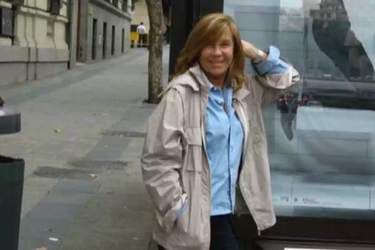 Mujer sonriente con chaqueta beige y camisa azul posando en una acera urbana junto a una parada de autobús Mujer sonriente con chaqueta beige y camisa azul posando en una acera urbana junto a una parada de autobús