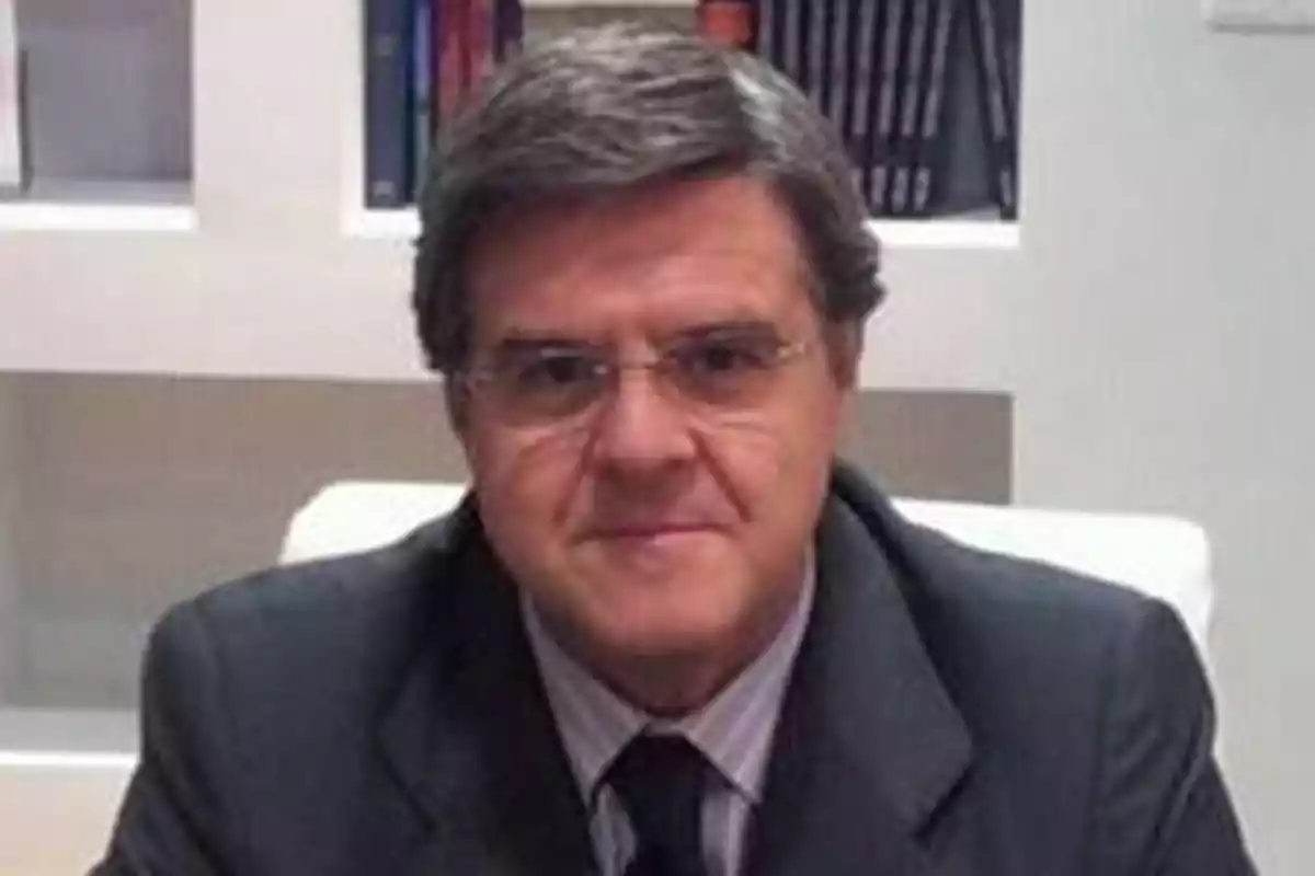 Hombre de cabello canoso y gafas con traje oscuro sentado frente a una estantería con libros Hombre de cabello canoso y gafas con traje oscuro sentado frente a una estantería con libros