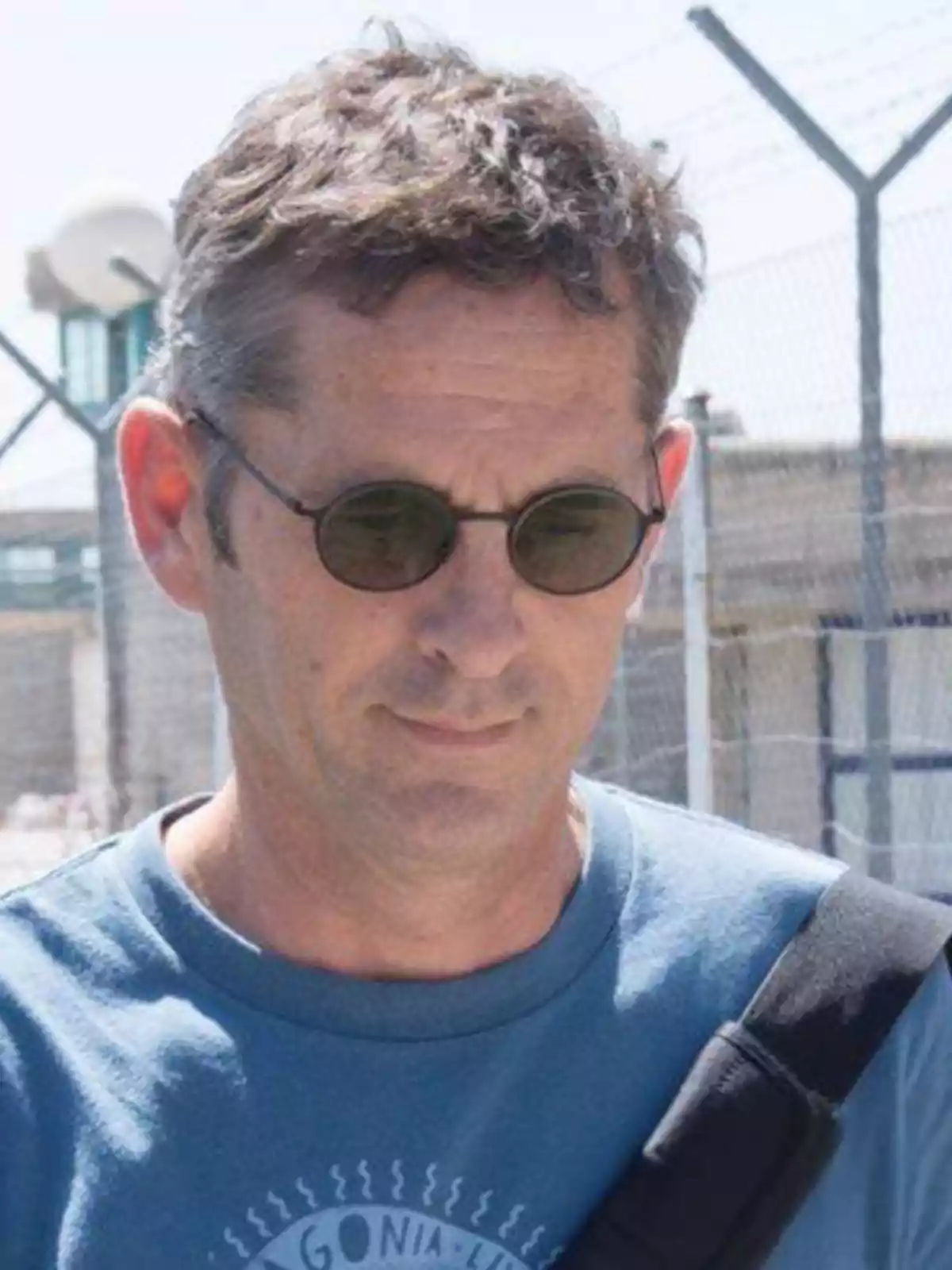 Hombre de cabello corto y gafas oscuras con camiseta azul y correa negra al hombro frente a una cerca de alambre Hombre de cabello corto y gafas oscuras con camiseta azul y correa negra al hombro frente a una cerca de alambre