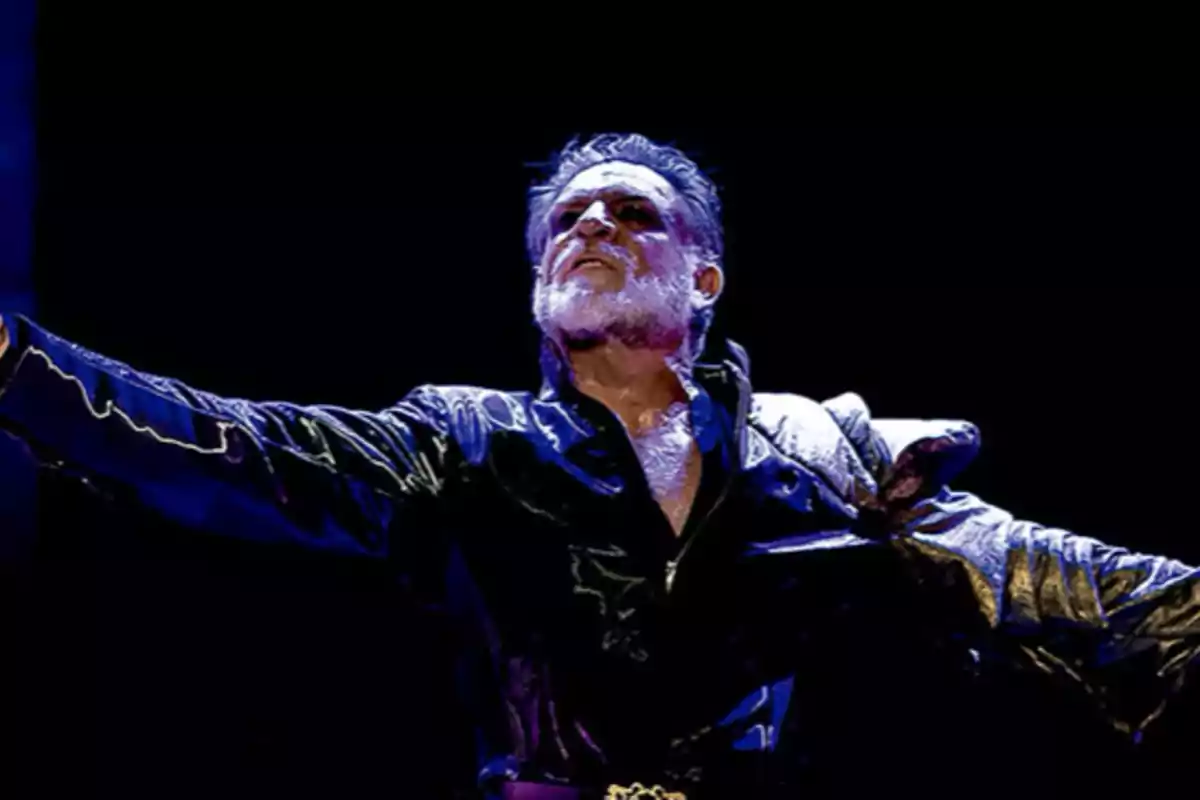Hombre con barba y cabello canoso vestido con un abrigo oscuro, brazos extendidos y expresión dramática en un escenario iluminado