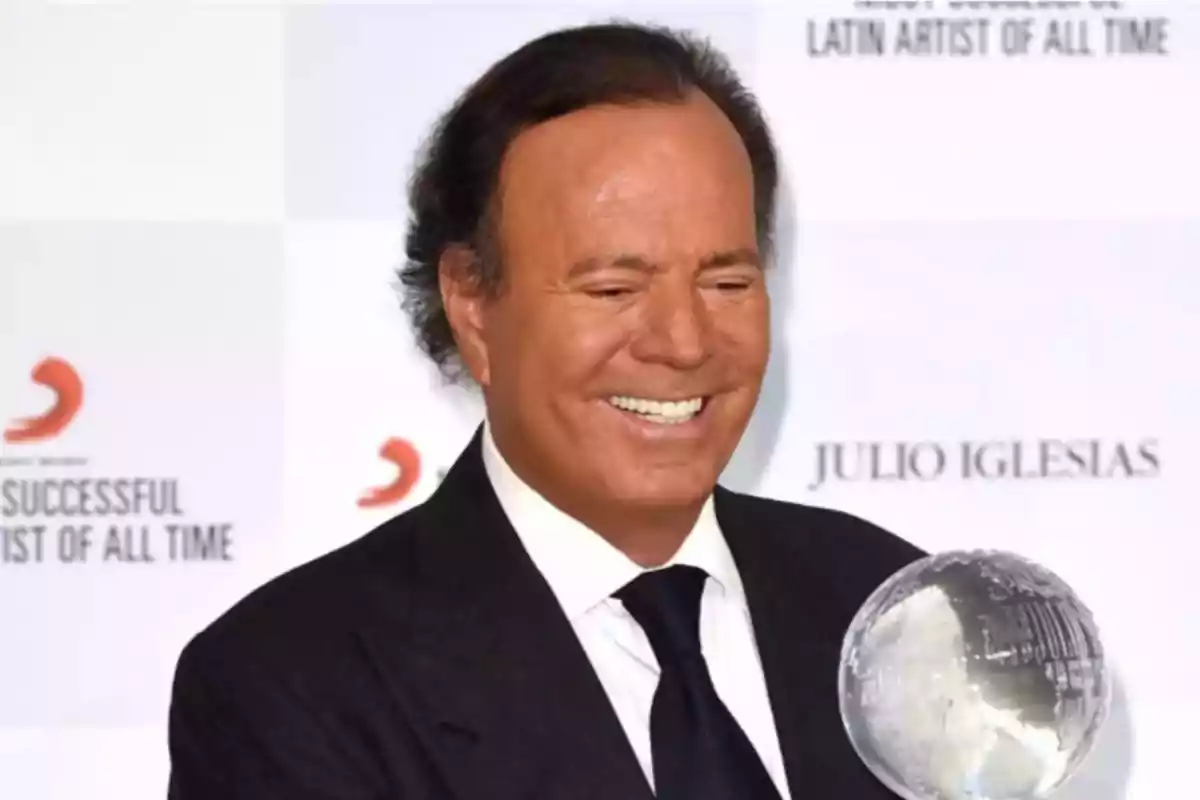 Hombre de traje oscuro sonriendo mientras sostiene un trofeo de cristal con fondo de pared blanca con texto borroso Hombre de traje oscuro sonriendo mientras sostiene un trofeo de cristal con fondo de pared blanca con texto borroso
