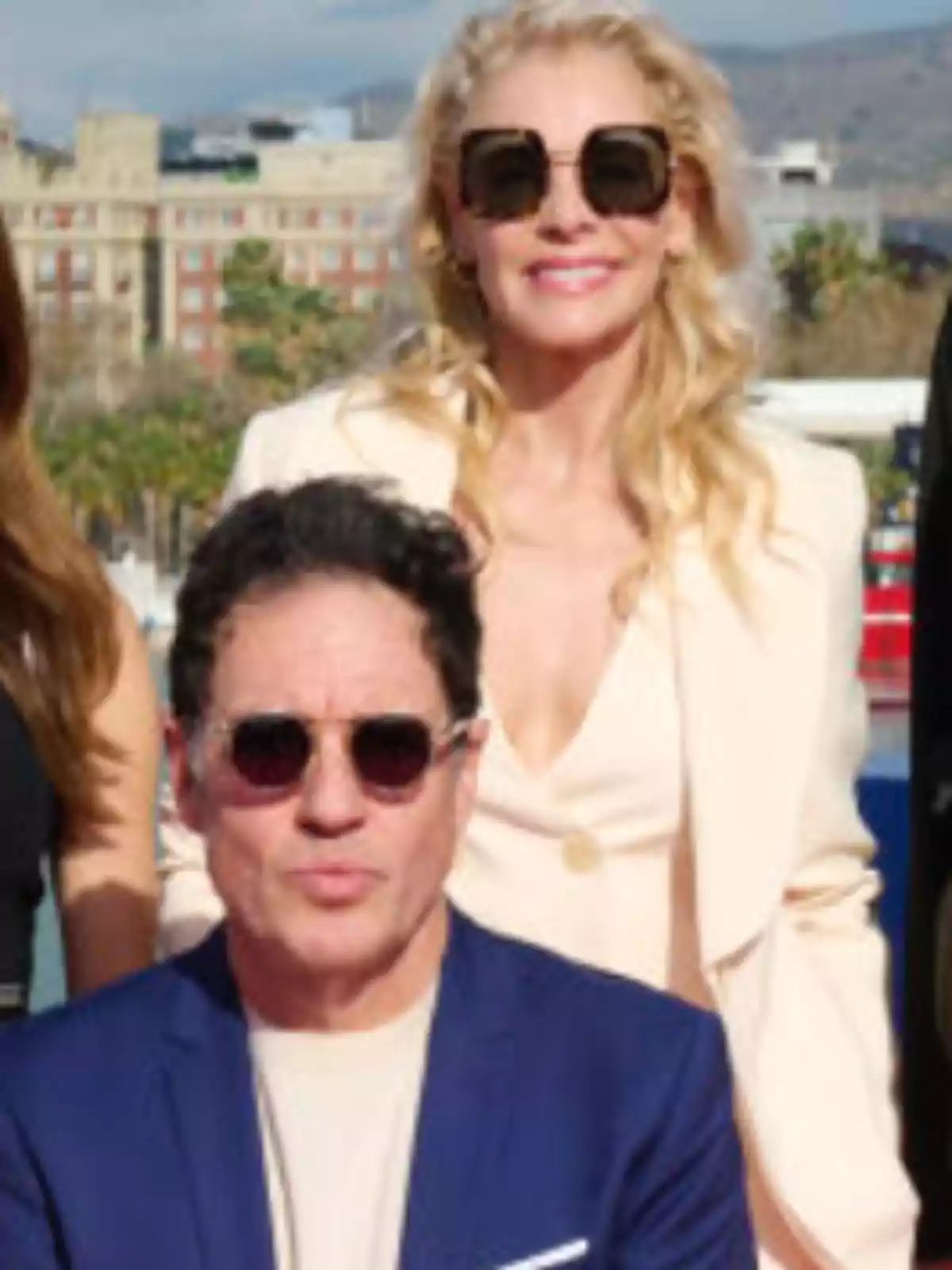 Dos personas posan al aire libre usando gafas de sol y ropa elegante, con un edificio y montañas al fondo Dos personas posan al aire libre usando gafas de sol y ropa elegante, con un edificio y montañas al fondo